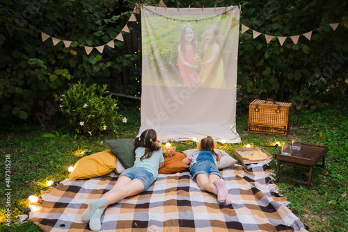 Family summer outdoor movie night. Girls lying on blanket and pillows, eating homemade popcorn and watching film on DIY screen with from projector. Summer outdoor weekend activities with kids.