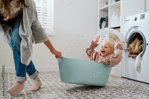 Mom is playing with daughter who is sitting in laundry bowl little girl wants to spend time with woman and help with household chores mother drags her daughter around laundry room for fun in bowl photo