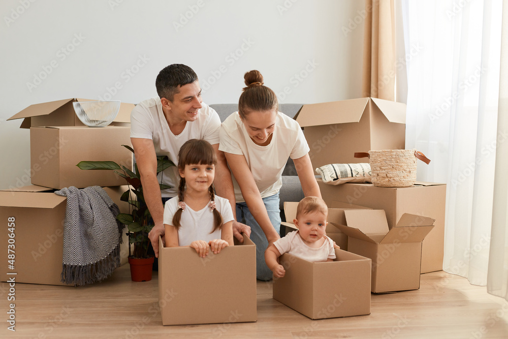 Happy positive Caucasian couple sitting on floor having fun together during moving to a new home, wearing white t-shirts, parents playing with their children in cardboard boxes. Happy positive Caucasian couple sitting on floor having fun together during moving to a new home, wearing white t-shirts, parents playing with their children in cardboard boxes.
