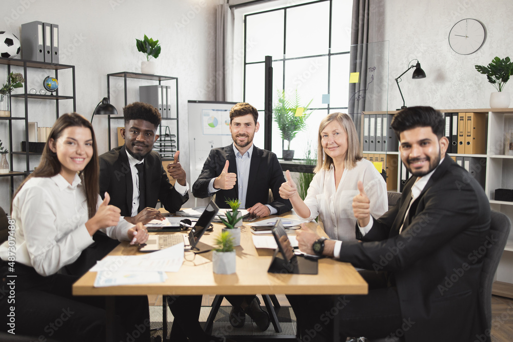 Team of five multiracial business people in formal clothes sitting together at office desk, smiling and looking at camera and showing thumb up. Conference meeting of diverse colleagues. Team of five multiracial business people in formal clothes sitting together at office desk, smiling and looking at camera and showing thumb up. Conference meeting of diverse colleagues.