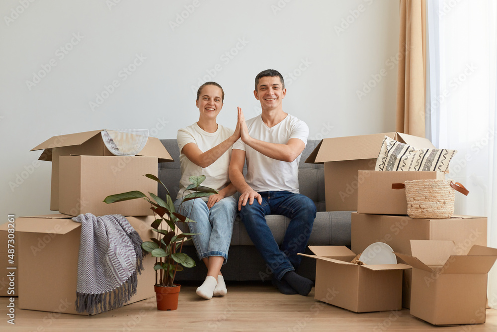 Indoor shot of young family buying new flat and posing surrounded with boxes with personal pile, sitting on sofa with palms together, looking at camera with happy facial expression. Indoor shot of young family buying new flat and posing surrounded with boxes with personal pile, sitting on sofa with palms together, looking at camera with happy facial expression.