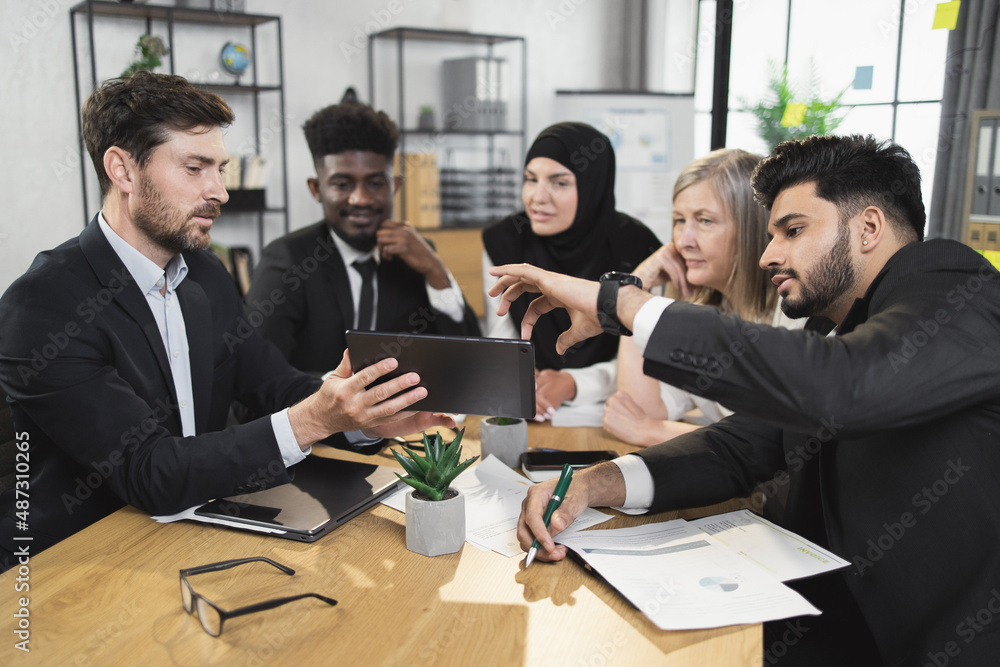 Multi ethnic team of business partners looking on one tablet screen during brainstorming at conference room. Five people discussing common working project at office. Multi ethnic team of business partners looking on one tablet screen during brainstorming at conference room. Five people discussing common working project at office.