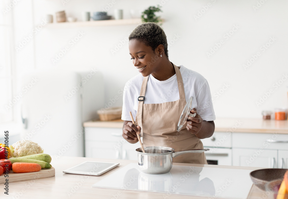 Glad young african american lady in apron cooking and watching video lesson on tablet Glad young african american lady in apron cooking and watching video lesson on tablet