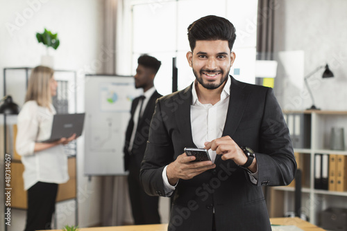 Portrait of indian businessman with smartphone in hands smiling on camera while his multiracial partners talking on background. Conference, teamwork and people concept.