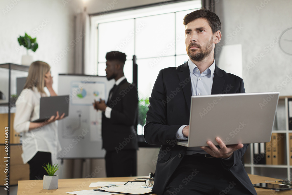 Caucasian man in suit using laptop and looking aside while leaning on desk at modern office. Multiracial company workers standing on background and talking. Caucasian man in suit using laptop and looking aside while leaning on desk at modern office. Multiracial company workers standing on background and talking.