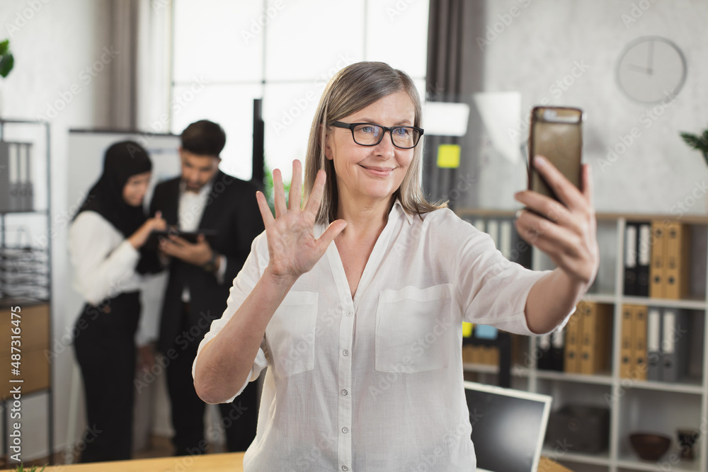Confident mature business lady in eyeglasses using smartphone for video conference at modern office. Blur background of two multiracial colleagues using digital tablet. Confident mature business lady in eyeglasses using smartphone for video conference at modern office. Blur background of two multiracial colleagues using digital tablet.