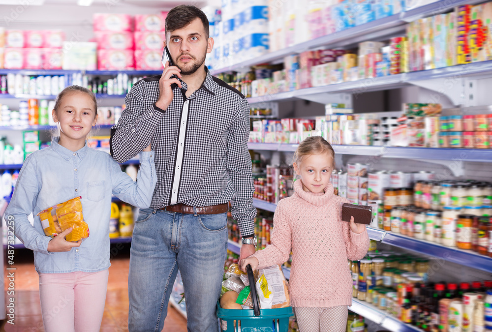 Young father speaking phone while doing shopping with daughters in supermarket