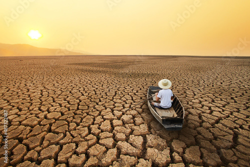 Young man paddle a boat at dry cracked earth with orange sky and hot weather of the sun. Metaphor Climate change, Drought and water crisis concept.