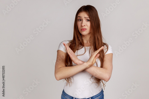 Portrait of upset disappointed girl crossing hands, showing stop denial gesture, saying definitive no, this is the end, wearing white T-shirt. Indoor studio shot isolated on gray background.