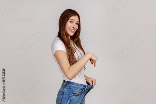 Portrait of happy attractive beautiful slim young woman showing successful weight loss, pointing at big jeans, wearing white T-shirt. Indoor studio shot isolated on gray background.