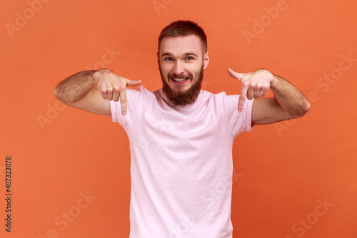 Cheerful satisfied man pointing fingers down, paying attention to advertising area, blogger recommending to subscribe, wearing pink T-shirt. Indoor studio shot isolated on orange background.