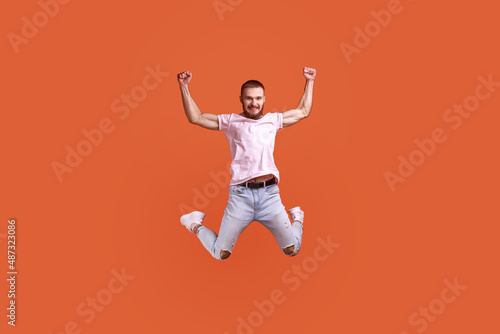 Portrait of happy attractive bearded man jumping in air raised arms, looking at camera, copy space for ad, wearing pink T-shirt and jeans. Indoor studio shot isolated on orange background.