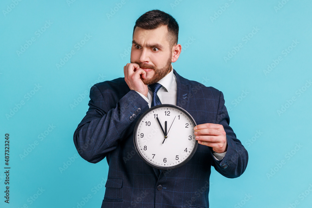 Portrait of impatient anxious businessman in dark suit holding big clock and biting nails, nervous about delayed meeting or deadline, need hurry up. Indoor studio shot isolated on blue background. Portrait of impatient anxious businessman in dark suit holding big clock and biting nails, nervous about delayed meeting or deadline, need hurry up. Indoor studio shot isolated on blue background.