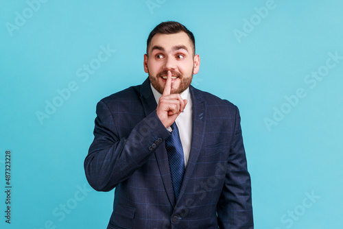 Portrait of businessman wearing official style suit holding finger near lips showing shh gesture, keeping secrets preparing surprise. Indoor studio shot isolated on blue background.