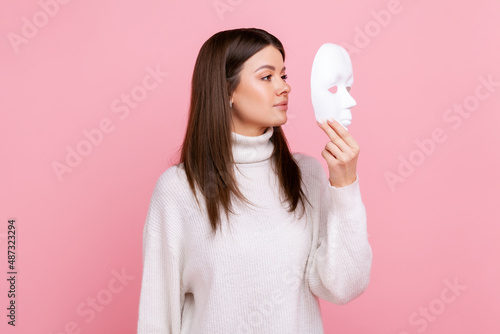 Side view portrait of girl holds white face mask, has mysterious expression, hiding his personality, wearing white casual style sweater. Indoor studio shot isolated on pink background. photo