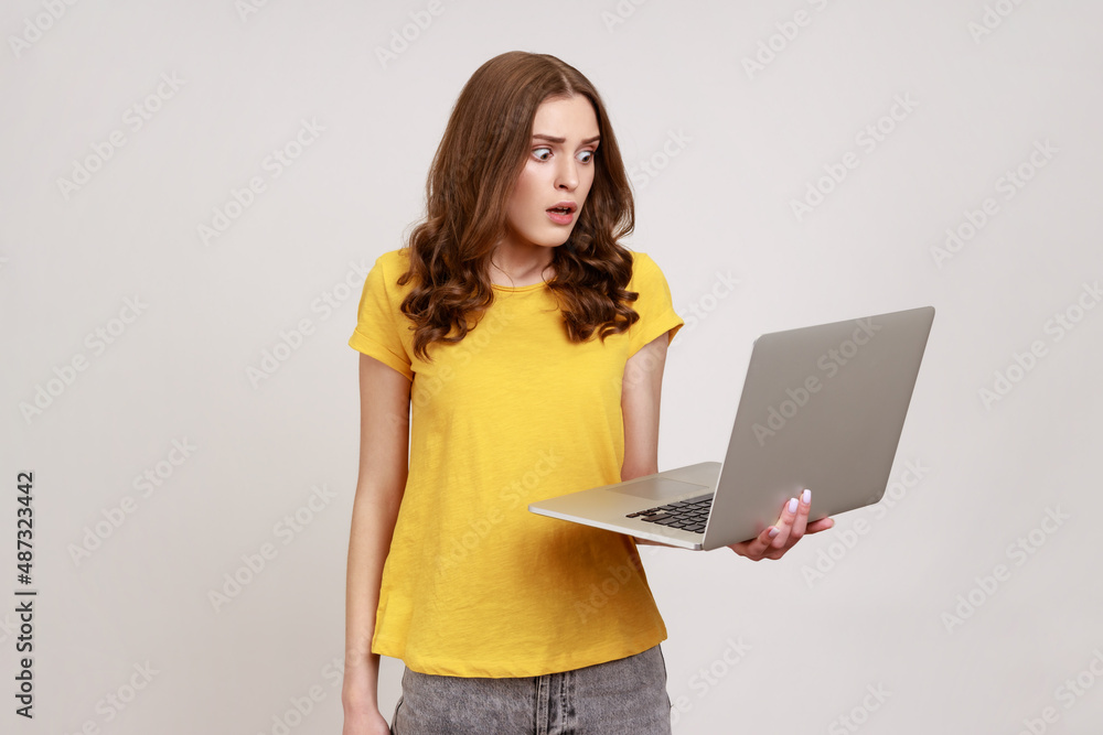 Naklejka premium Portrait of scared freelancer working on computer. Amazed young woman in casual T-shirt reading shocking news on laptop, working online using computer. Indoor studio shot isolated on gray background.