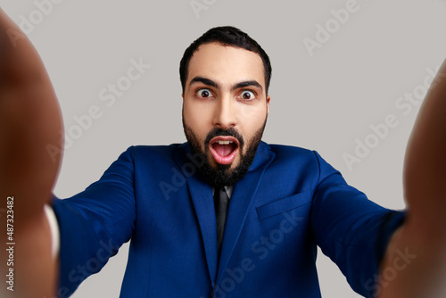 Astonished bearded young adult man taking selfie, looking at camera with shocked expression and open mouth POV, wearing official style suit. Indoor studio shot isolated on gray background. © khosrork