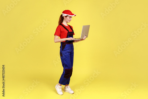 Side view portrait of smiling worker woman working on laptop, processes online orders, expressing positive emotions, wearing overalls and red cap. Indoor studio shot isolated on yellow background.