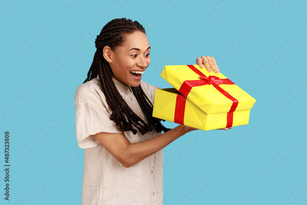 Portrait of happy excited woman with black dreadlocks looking into gift box, opening present and peeking inside with happiness, wearing white shirt. Indoor studio shot isolated on blue background. Portrait of happy excited woman with black dreadlocks looking into gift box, opening present and peeking inside with happiness, wearing white shirt. Indoor studio shot isolated on blue background.
