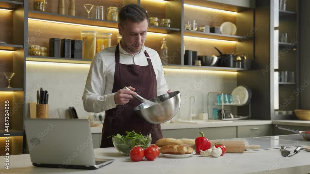 Young man in apron prepares dinner following video. Chef cooks dish with vegetables mixing sauce in bowl and watching recipe in laptop at table in kitchen Young man in apron prepares dinner following video. Chef cooks dish with vegetables mixing sauce in bowl and watching recipe in laptop at table in kitchen
