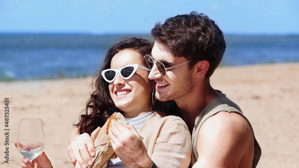 Cheerful man in sunglasses holding baguette and hugging girlfriend with wine on beach