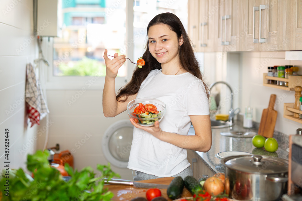 Portrait of young positive woman eating healthy vegetable salad at kitchen