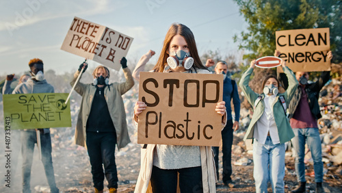Portrait of Attractive Young Woman Activist Holding a Poster Calling to Stop Plastic. In Background Fighting People Protesting Against Garbage Pollution Staying at Dump in City Outskirts. © alex_marina