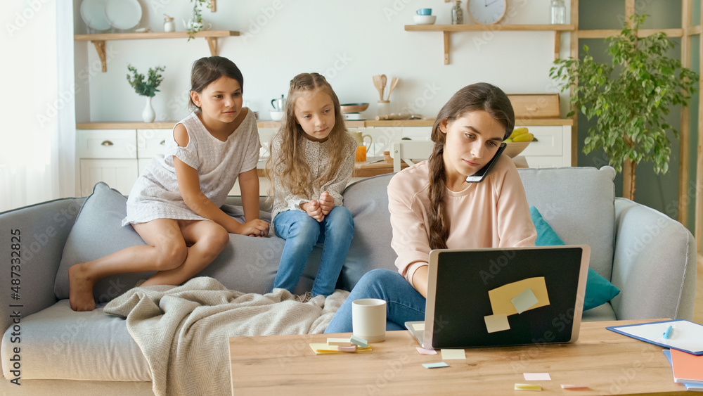 A Busy Mother Working Remotely With Her Children's at Home. The Daughters are Sits on the Couch, While Their Mother Working With a Laptop at the Table and Doing Business Conversation on the Phone. A Busy Mother Working Remotely With Her Children's at Home. The Daughters are Sits on the Couch, While Their Mother Working With a Laptop at the Table and Doing Business Conversation on the Phone.