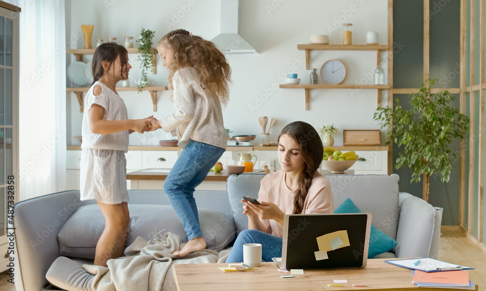 A Busy Mother Trying to Work Remotely at Home. On The Background Her Daughters are Running Around The Table, While Their Mother Working With a Laptop and Doing Business Conversation on the Phone. A Busy Mother Trying to Work Remotely at Home. On The Background Her Daughters are Running Around The Table, While Their Mother Working With a Laptop and Doing Business Conversation on the Phone.
