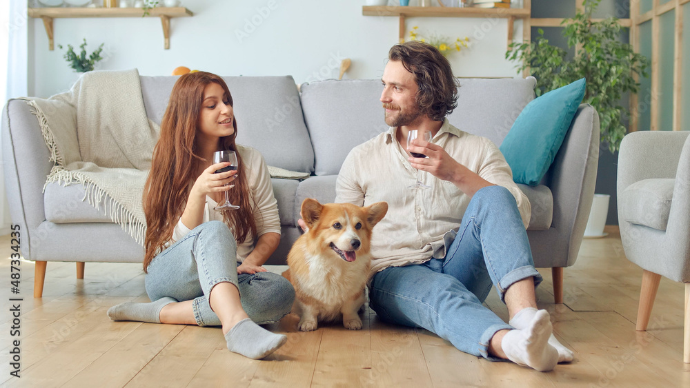 Happy Young Married Couple Sitting on the Floor Near a Couch and Clinking Glasses With Wine, celebrating new house purchase, successful mortgage investment. Cute Dog Sitting Between Married Couple.