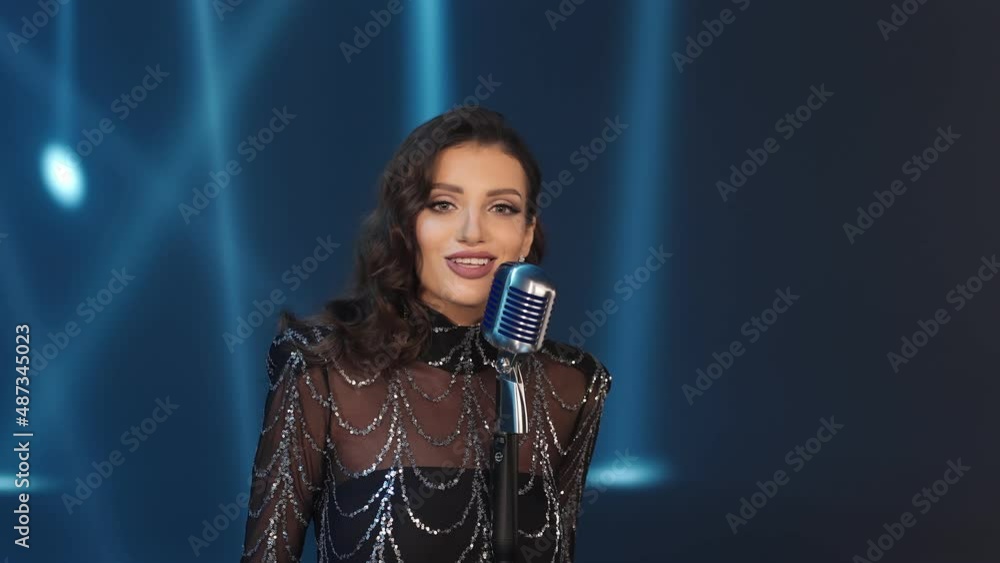 Beautiful Caucasian girl singing into a microphone on a blue background. European woman singer on the spotlight. Shooting a video. Brunette in a shiny evening dress. Music band. Beautiful Caucasian girl singing into a microphone on a blue background. European woman singer on the spotlight. Shooting a video. Brunette in a shiny evening dress. Music band.