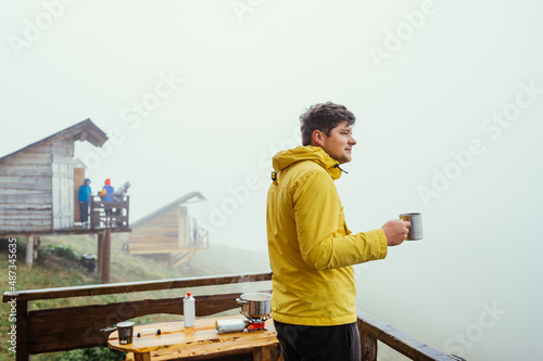 Male hiker in a yellow jacket stands on the balcony of a house in the mountains with a cup of hot drink in his hands on a background of foggy views.
