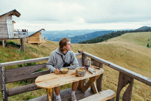 An attractive man sits on the balcony of a mountain house at a table with food and looks away with a serious face.
