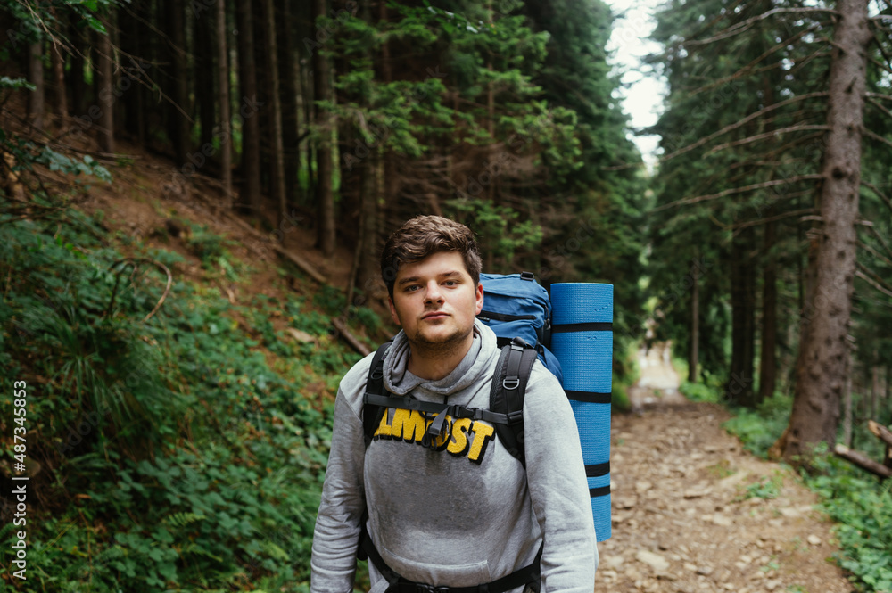 Male hiker with a backpack on his back walking on a trail in the mountains and climbing up, looking at the camera with a serious face.