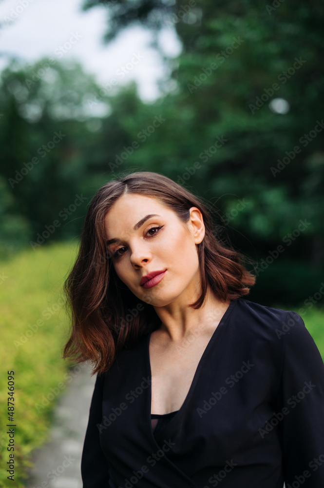 Stylish brunette woman in a jacket stands in the park and looks at the camera on the background of the park, vertical.