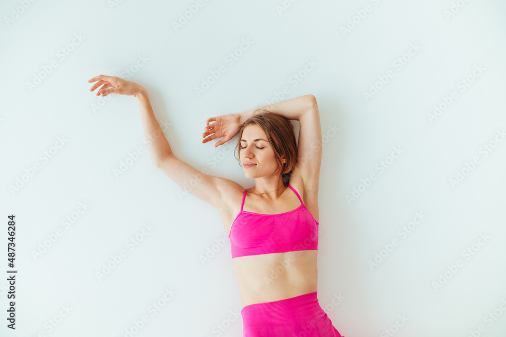 Positive sports woman in pink sportswear lies on a white background with closed eyes and smiles.