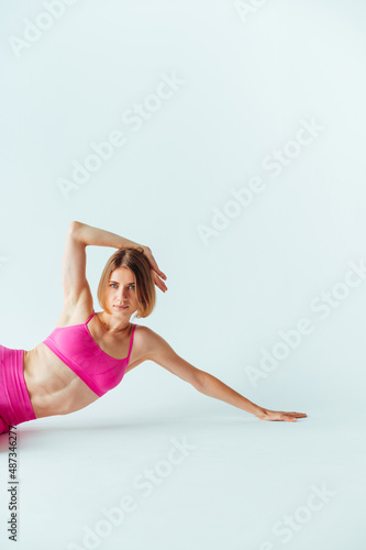 Beautiful sporty woman trains on a white background in pink clothes, lying on the ground and posing for the camera. Vertical