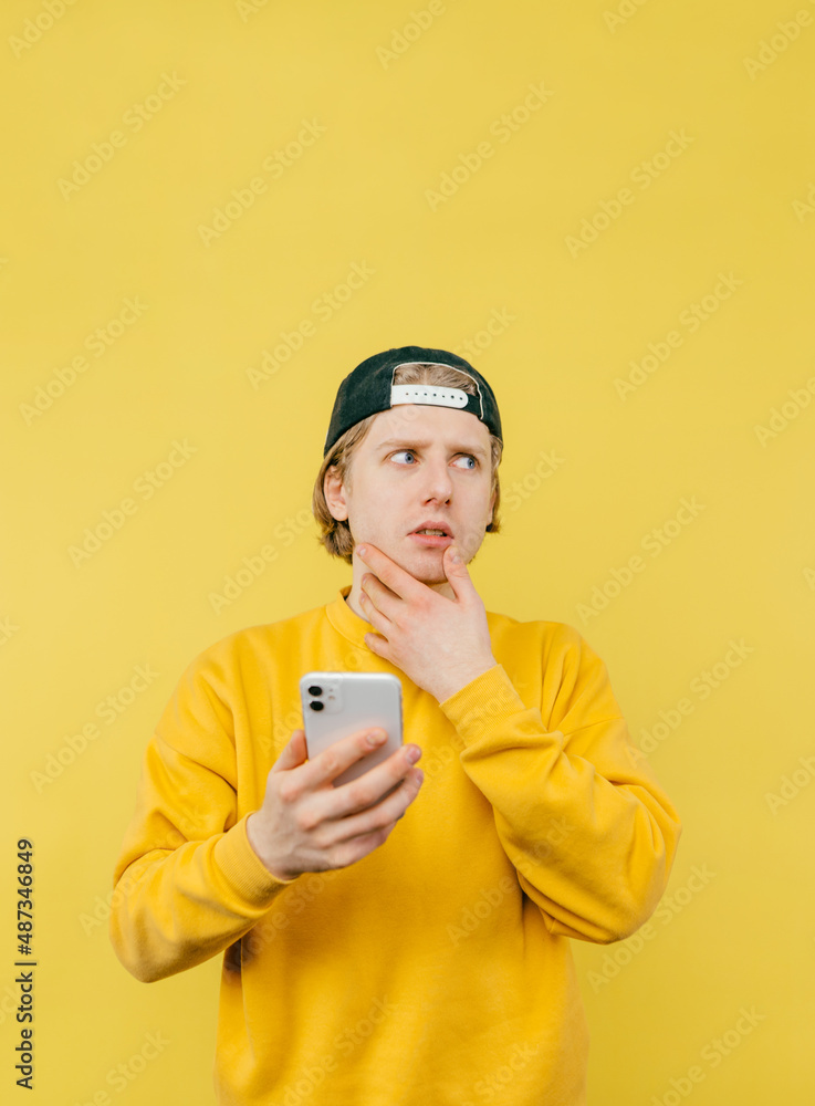 Portrait of a pensive guy in a sweatshirt and cap on a yellow background with a smartphone in his hand scratches his beard and looks away. Vertical