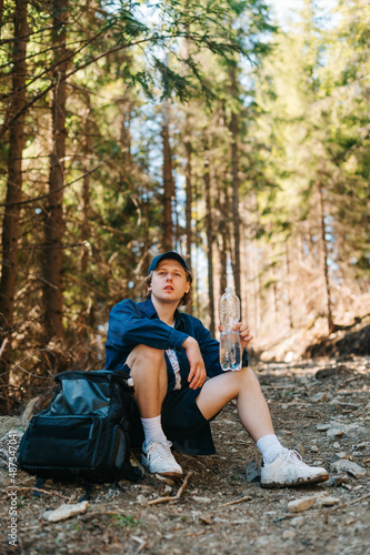 Young tourist sits on a trail in the mountains through the woods with a backpack and drinks water from a bottle and rests.
