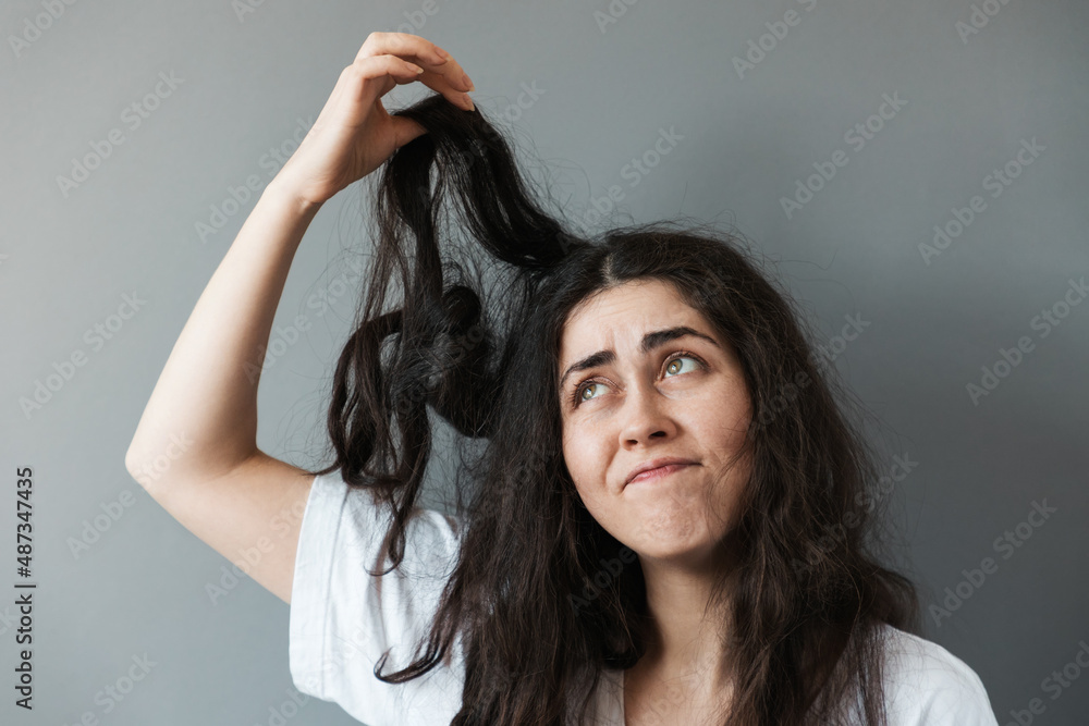 A young disgruntled woman looks at the tips of her disheveled long dark hair, pulling a stand up. Gray background. The concept of hair problems A young disgruntled woman looks at the tips of her disheveled long dark hair, pulling a stand up. Gray background. The concept of hair problems
