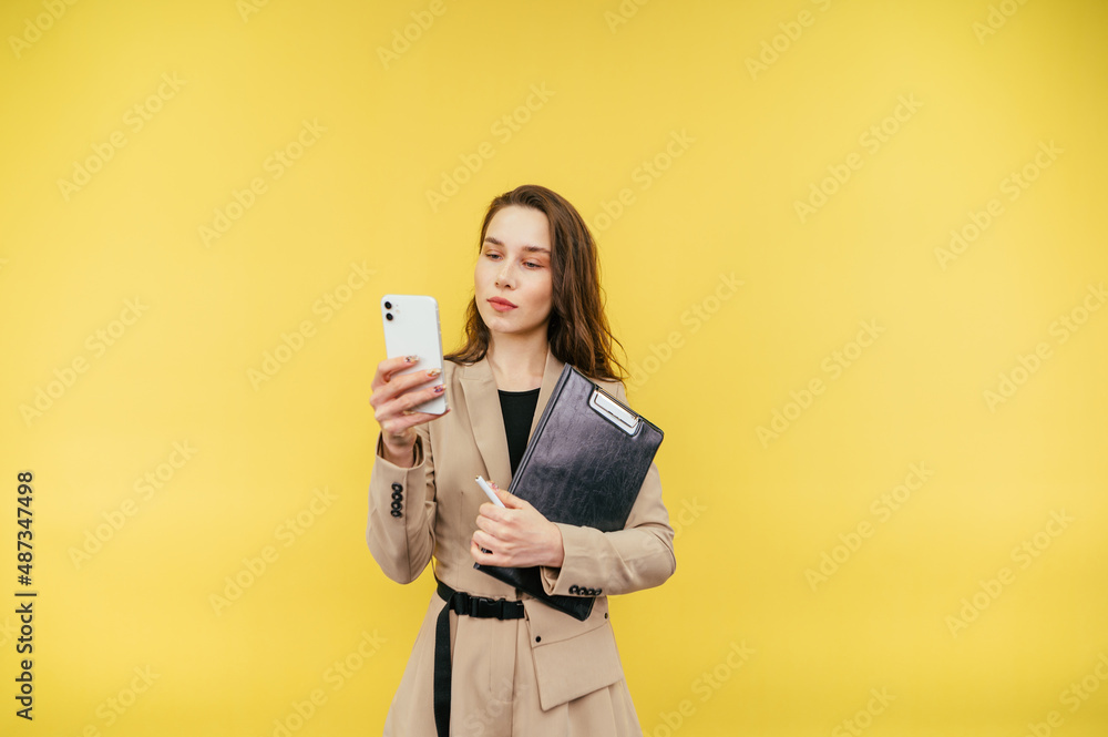 Attractive business woman in a jacket with a tablet stands on a yellow background and uses a smartphone with a serious face. Attractive business woman in a jacket with a tablet stands on a yellow background and uses a smartphone with a serious face.