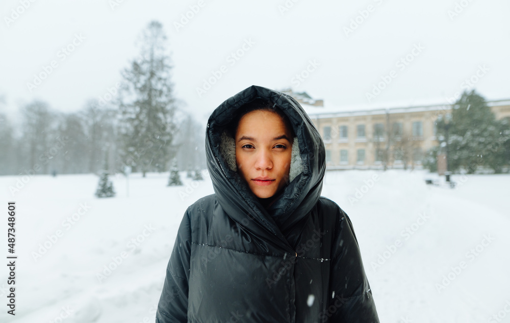 Beautiful hispanic woman in a black coat and hood stands on the street in the snow and looks at the camera with a serious face. A lady in warm clothes walks down the street in winter. Beautiful hispanic woman in a black coat and hood stands on the street in the snow and looks at the camera with a serious face. A lady in warm clothes walks down the street in winter.