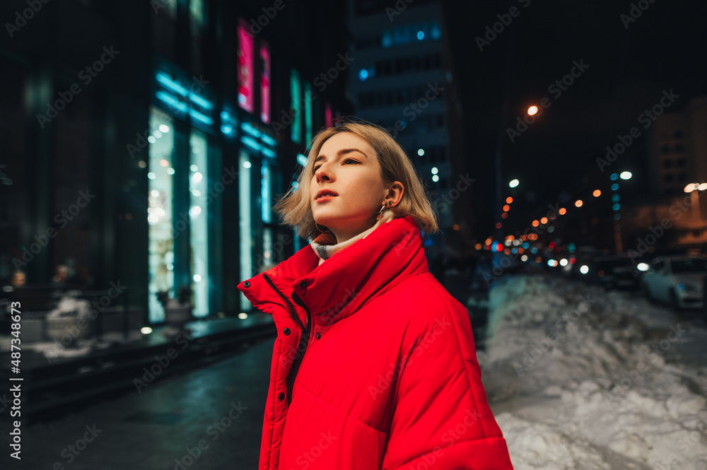 Fototapeta premium Portrait of a beautiful woman in a red jacket on the street at night, looking aside against the backdrop of the city evening landscape