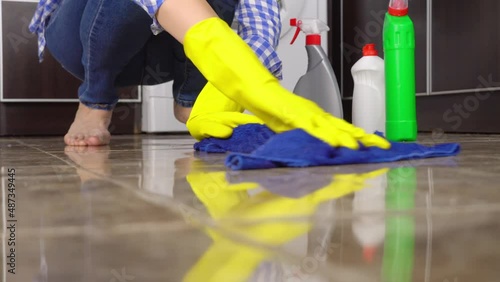 young woman in close-up washes squatting or kneeling on floor. She runs blue rag across floor, collecting debris and dust. Yellow gloves. There are cleaning and cleaning products in background. photo