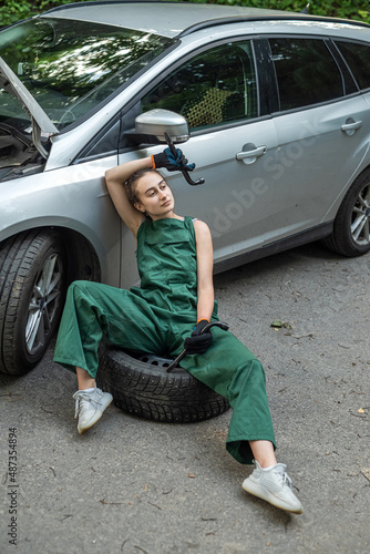 a female mechanic in overalls is resting near a broken car and waiting for help from a partner © RomanR