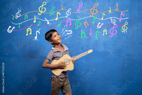 happy kid enjoys playing with cardboard guitar with musical signs on wall - concept of entertainment, poverty, childhood dream for rockstar.