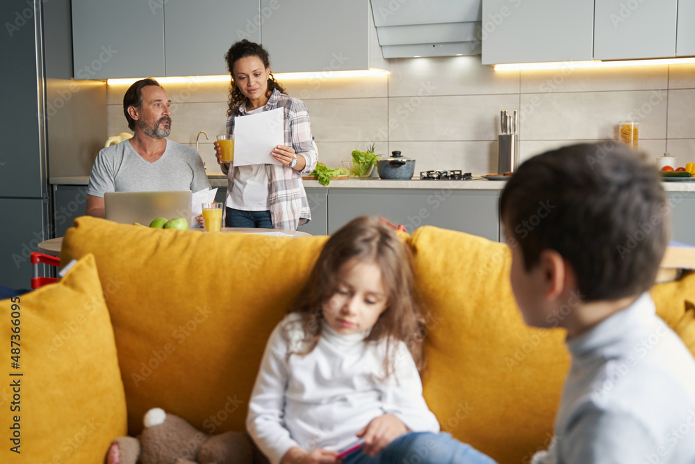 Female checking paper near husband with kids on sofa Female checking paper near husband with kids on sofa