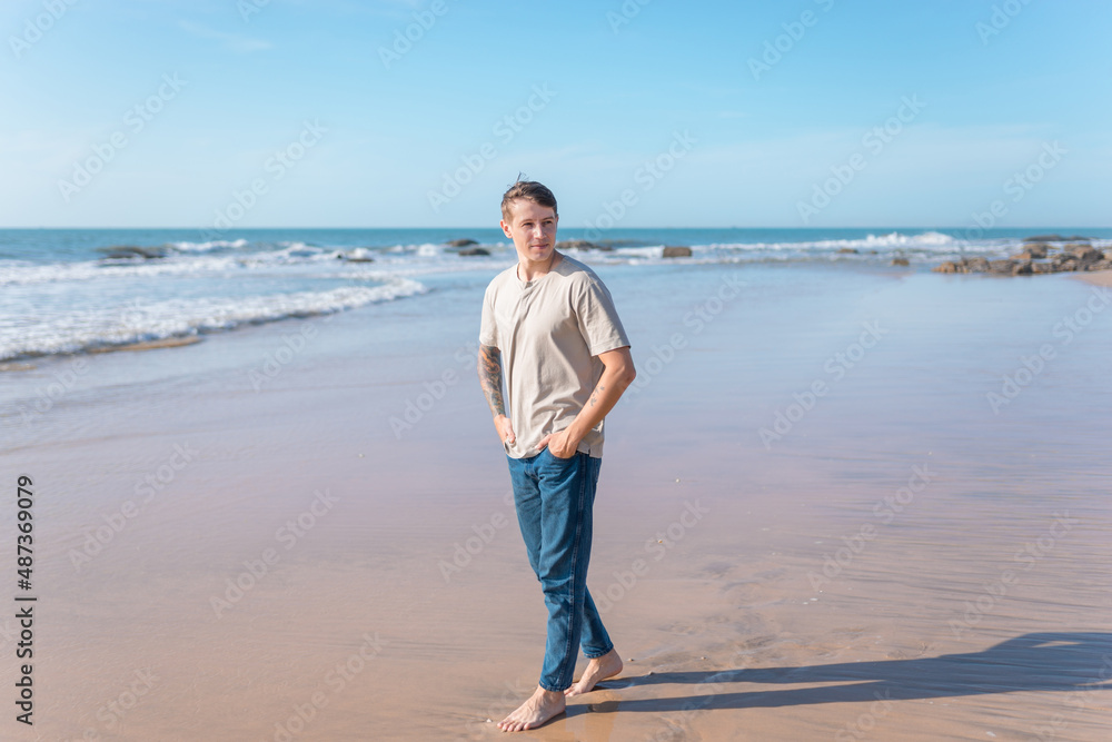 young caucasian man with full-arm tattoo in casual clothing walking on the beach