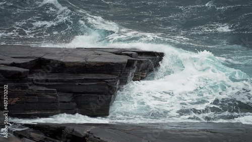 Heavy waves hit the dark jagged rocks on the shoreline. Turquoise water foaming and raging. Slow-morion. photo