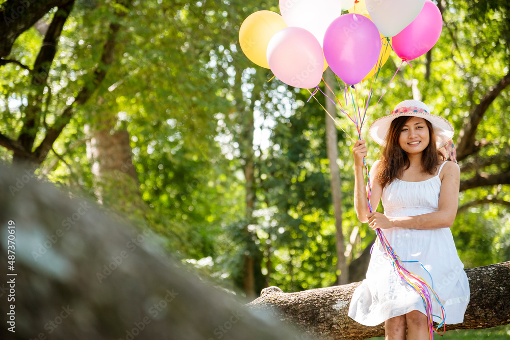 Cheerful beauty woman holding balloons relax sitting under big tree in green park with happiness. Woman Hands holding vibrant air balloons play on birthday party happy time summer on sunshine outdoor